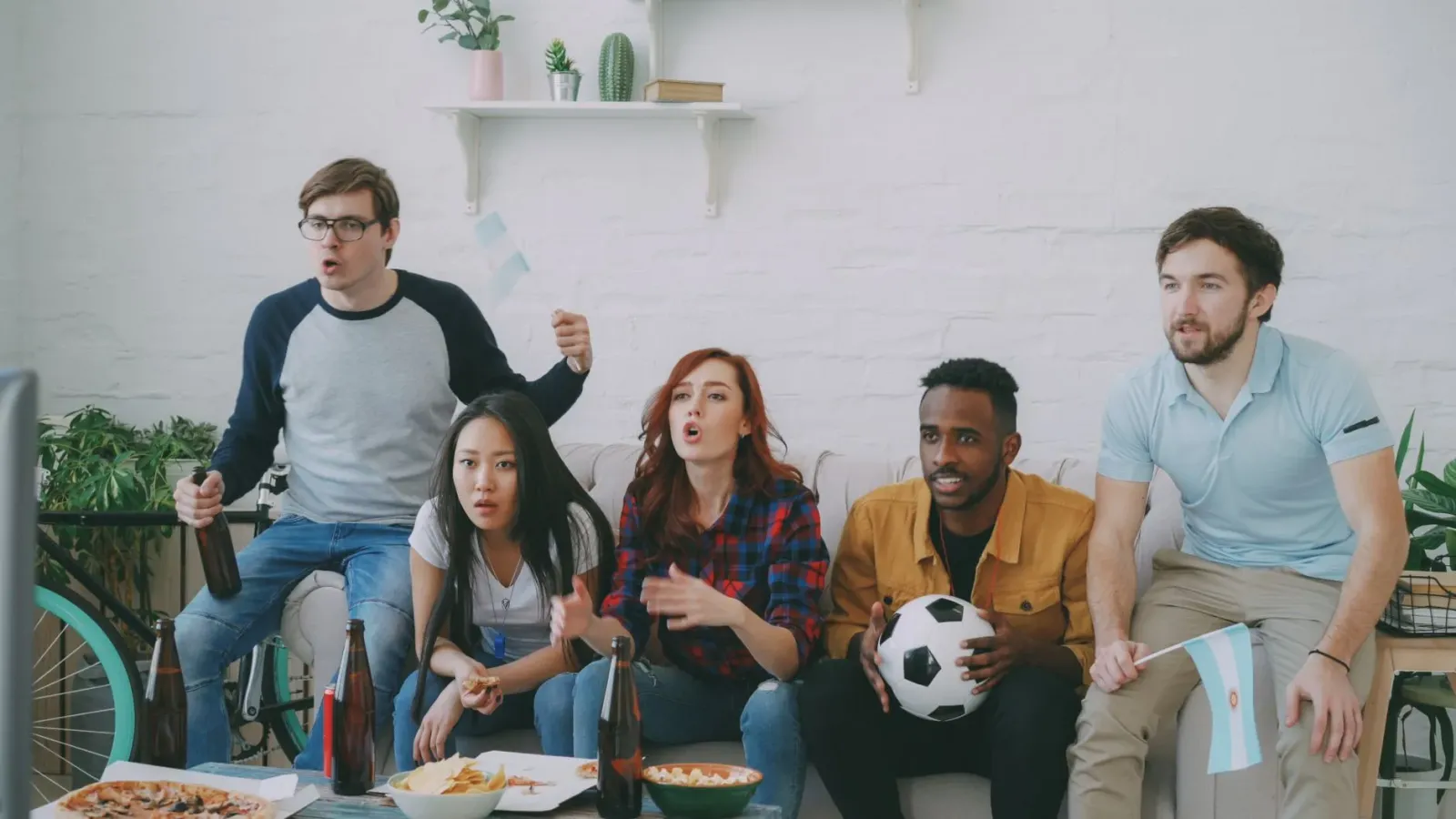 Fã sira hamutuk iha sala haree jogu UEFA Champions League iha TV boot, ho snacks iha meza, sorrisu no konsentrasaun