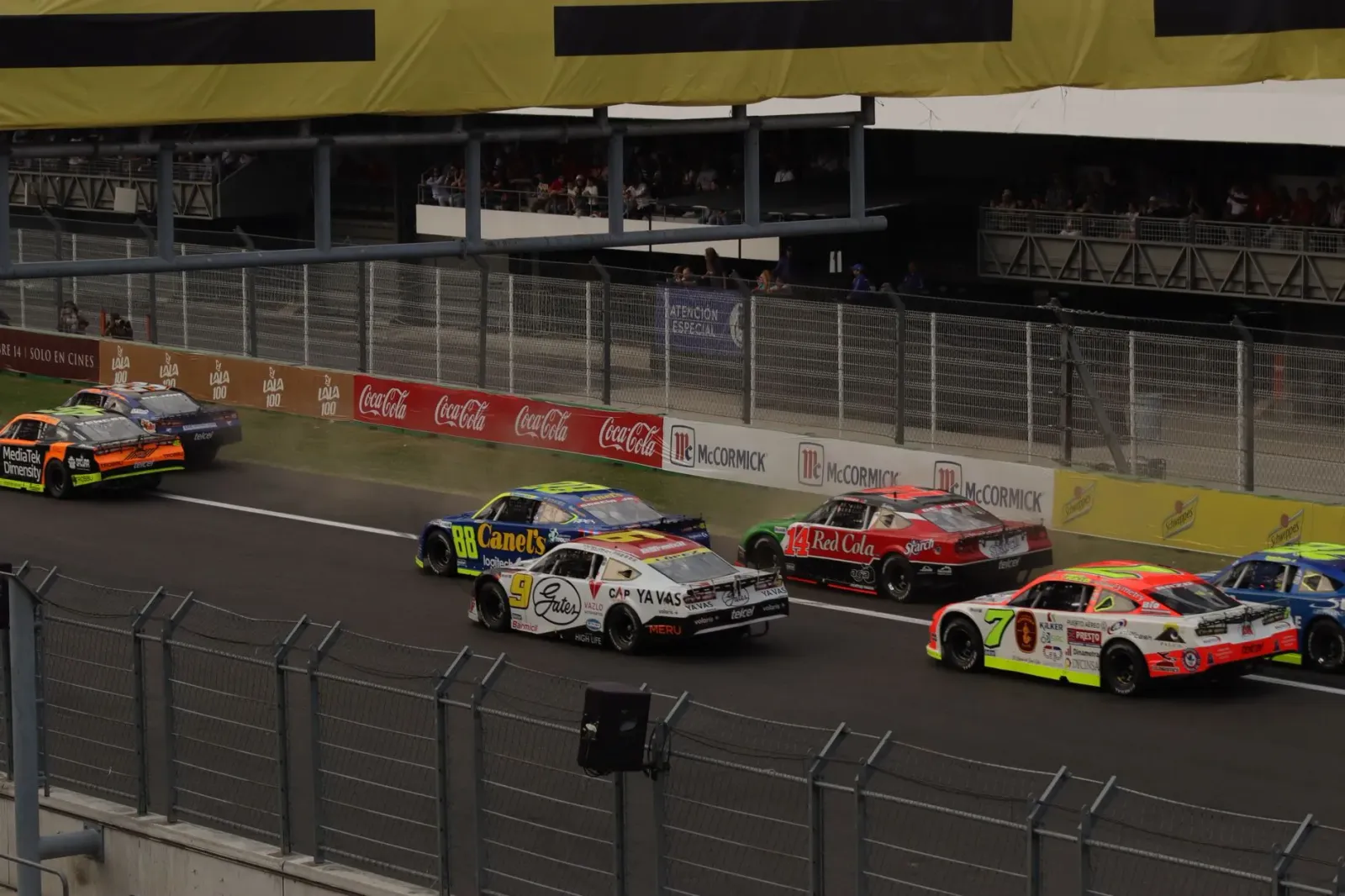 Imajen husi karru NASCAR ne'ebé kompeti iha pista, ho bandeira no ema barak iha arquibankada, hatudu ambiente kompetisaun des