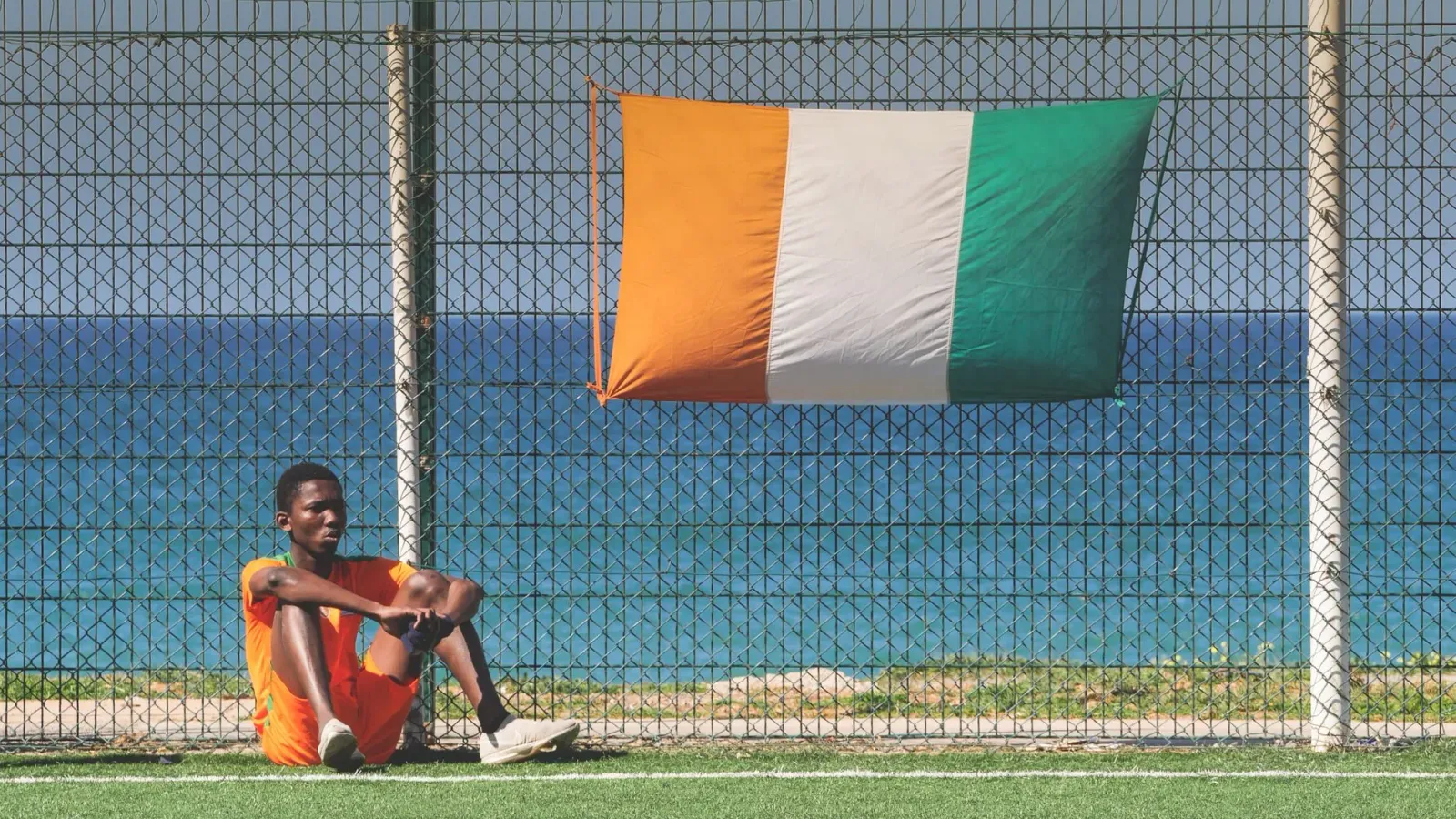 Imajen ida ne'ebe hatudu jogador futebol mane ida ho uniforme, iha fundu estadiu ho bandeira Ivory Coast, simboliza kazu inve