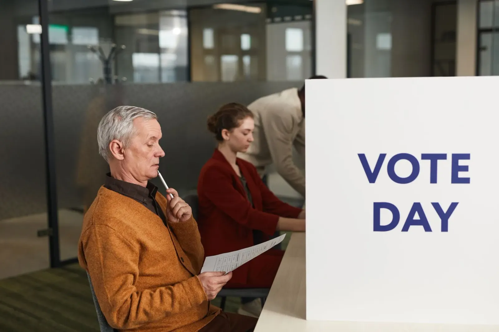 Imajen ida hatudu ema balu hela iha fila votasaun iha by-election, ho plaka sira no tenda informasaun, iha background vizinha