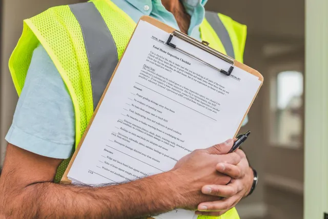 Imajen hatudu inspektor saúde halo verifikasaun iha edifísiu ho checklist no ekipamentu seguransa ahi, hanesan ezemplu ba pre