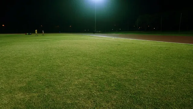 Imajen parke futebol iha Nova Iorke ho luz kalan, ema barak jogu hamutuk iha kampu verde, ho edifísiu sidade iha kotuk, atmos