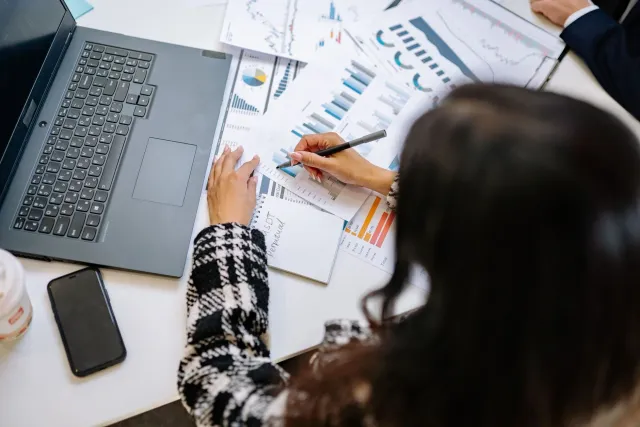 Imajen ida hatudu ema ida ho roupa formal haree dokumentu finanseiru no kalkuladór iha meza, ho naran KPMG no gráfiku iha kot