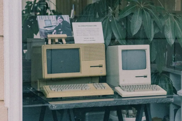 Ema ida tuur iha meja ho dokumentu no laptop, ho livru horóskopu no kotsaun Steve Jobs iha parede, hanesan ilustrasaun ba kon