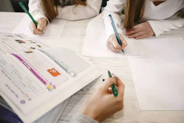 Imajen hatudu estudante sira uza papel no lapizeira iha sala aula, ho komputador iha fundu, ne'ebé hatudu balansu entre tekno