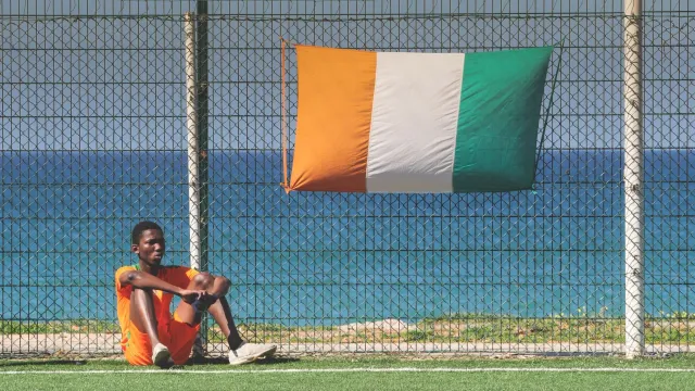 Imajen ida ne'ebe hatudu jogador futebol mane ida ho uniforme, iha fundu estadiu ho bandeira Ivory Coast, simboliza kazu inve