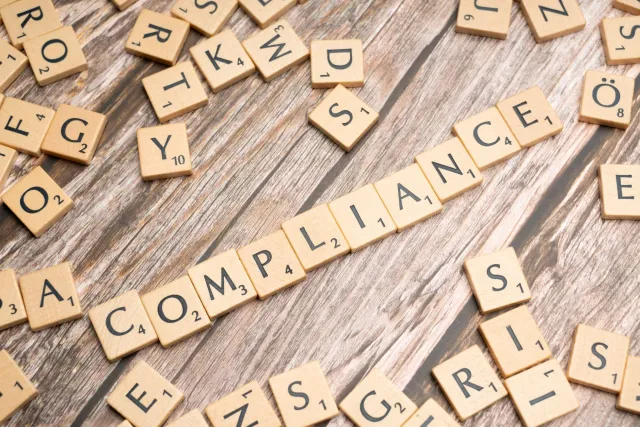 Imajen ida hatudu dokumentu compliance no relatóriu finanseiru iha meja, ho background kompanyas boot sira nia edifísiu. Símb