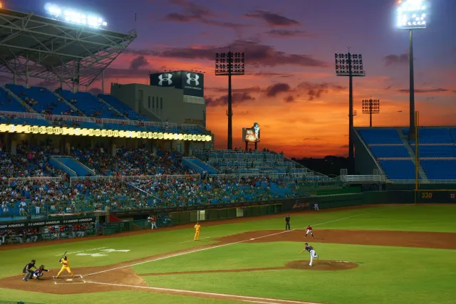 Imajen husi estadiu baseball ho ema barak iha kadeira, ho letra 'Faith & Family Night' iha monitor boot, iha kalan ho luz neo