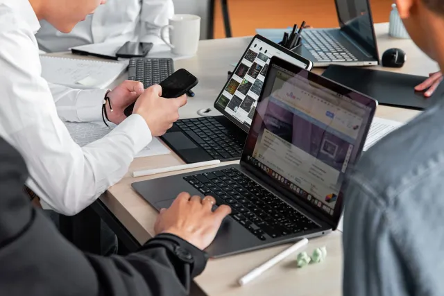 Imajen boot ne'ebé hatudu grupo ema iha reunisaun negósiu ho laptop no smartphone, iha foho iha fundu, hanesan ne'ebé mos iha