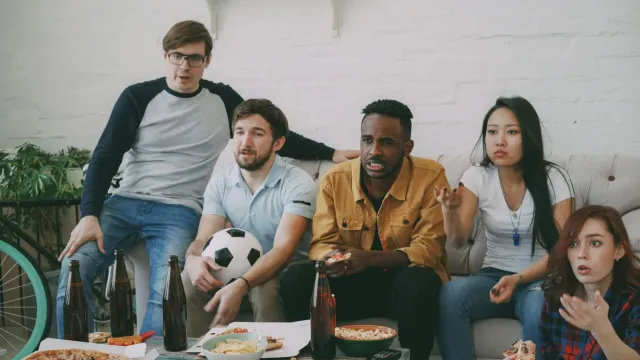 Imajen hatudu fã desportu barak hela iha sala, haree televizaun ne'ebé hatudu jogu futebol FIFA World Cup, ho emosaun no kons