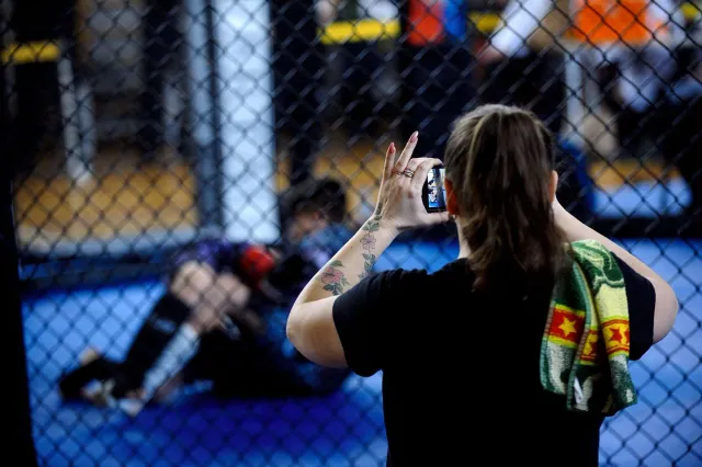 Imajen ida hatudu Cris Cyborg, kampiaun lendária MMA, iha okos lus brilhante, hamriik iha okos PFL cage, ho ninia rosto seriu