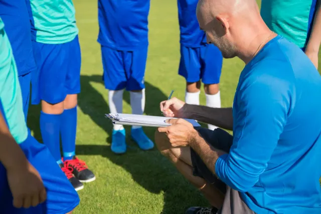 Imajen ida hatudu jogador sira iha kampu desportu servisu hamutuk ho treinador sira, ne'ebé reprezenta estratéjia longo prazu